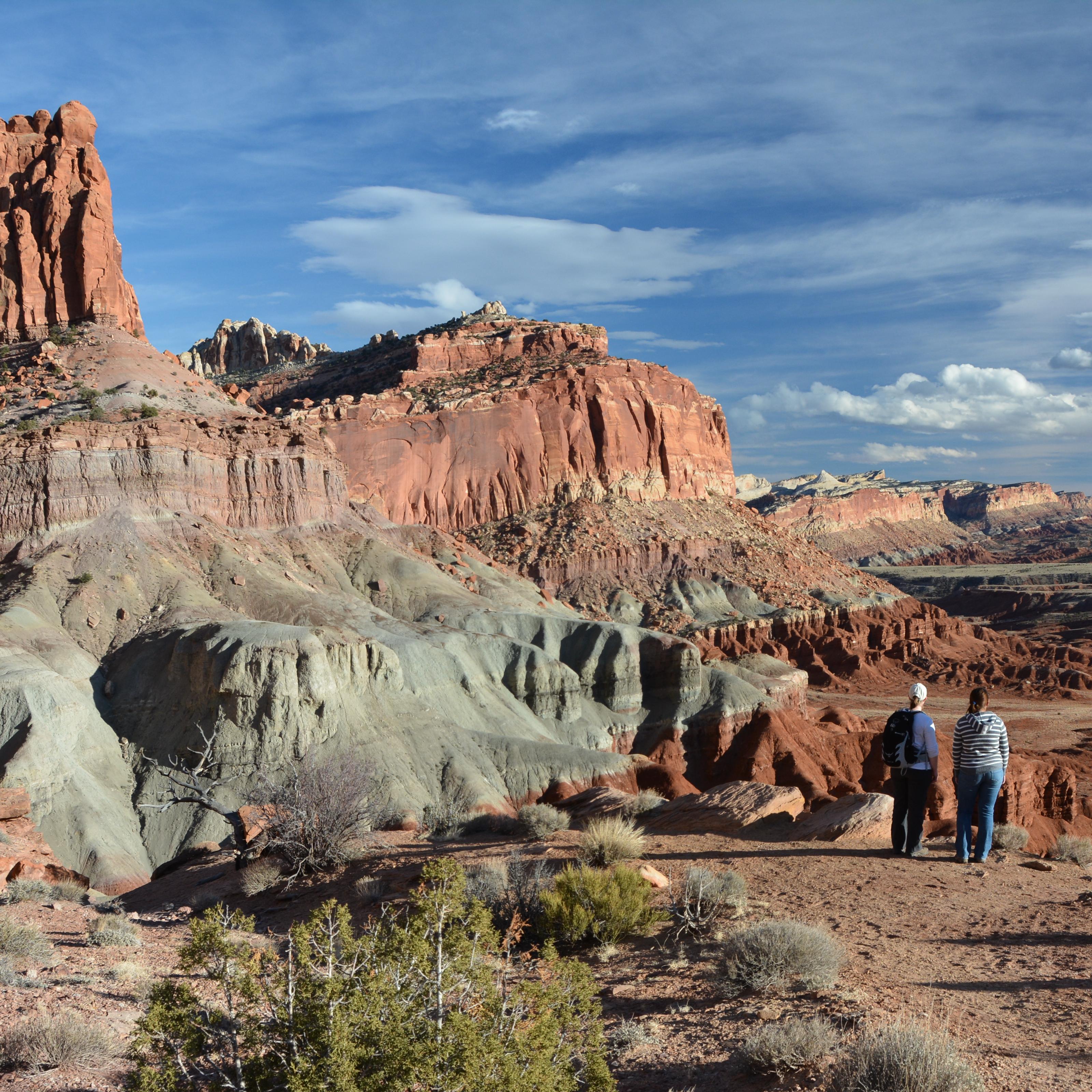 Two hikers stand at the edge of a cliff, looking at a panoramic view of colorful rock cliffs.