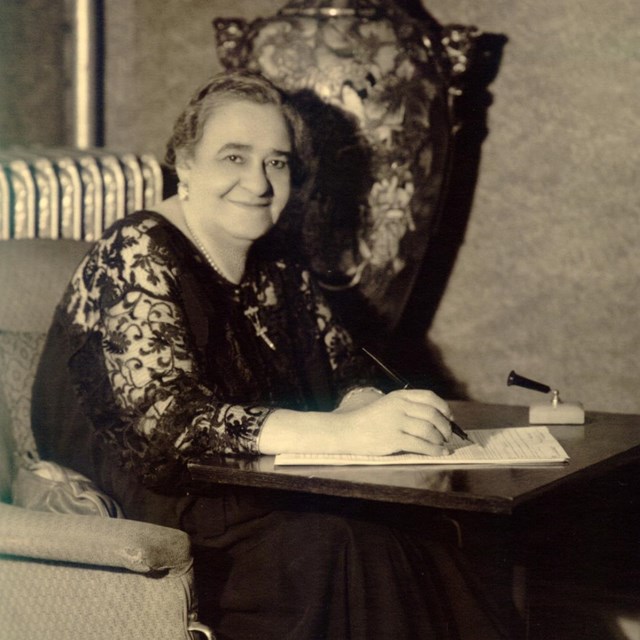 Black and white of an older woman sitting in a dark dress. She leans over a surface to write.