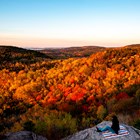 A person sits on a rock ledge looking out over sunny hills with colorful fall foliage
