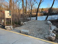 A gravel path lined with large rocks and trees leads to the river. To the left is an information pan