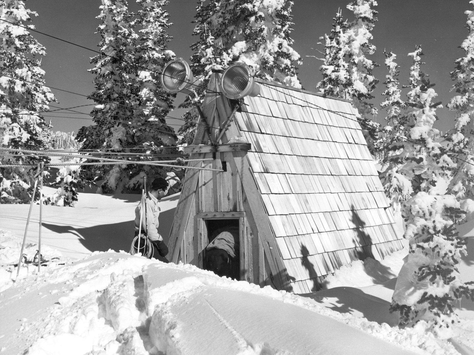 A steep A-frame roof building sits in deep snow surrounded by trees
