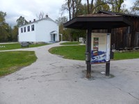 A branching walkway leads from a three sided kiosk past a brown barn to a two story white building.