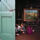A group of children sitting inside of a building on the carpet in front of a painting.