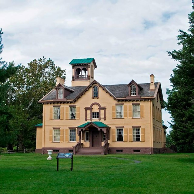 Large yellow plaster house with a green tower on it. It sits in a lush green clearing. 