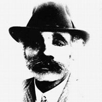 A black and white portrait of Mr. Fletcher. He wears a brimmed hat, suit and tie, and has a mustache
