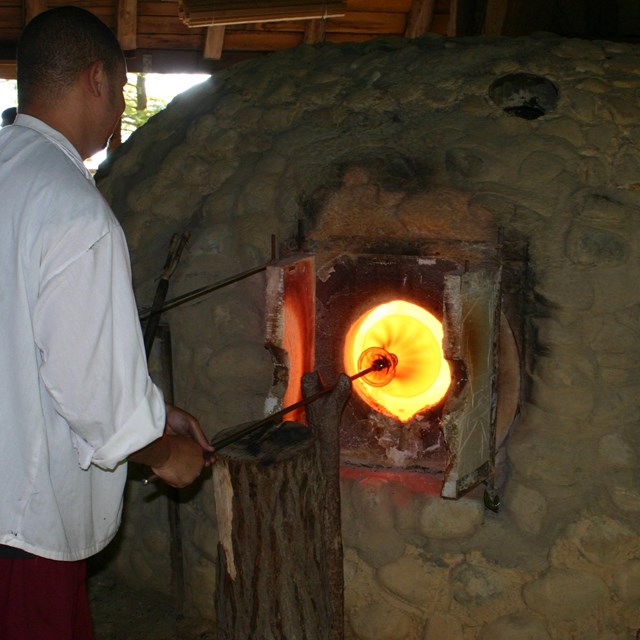 A yellow-orange sphere of glass is being heated in a brown stone kiln by a man in plain clothes.