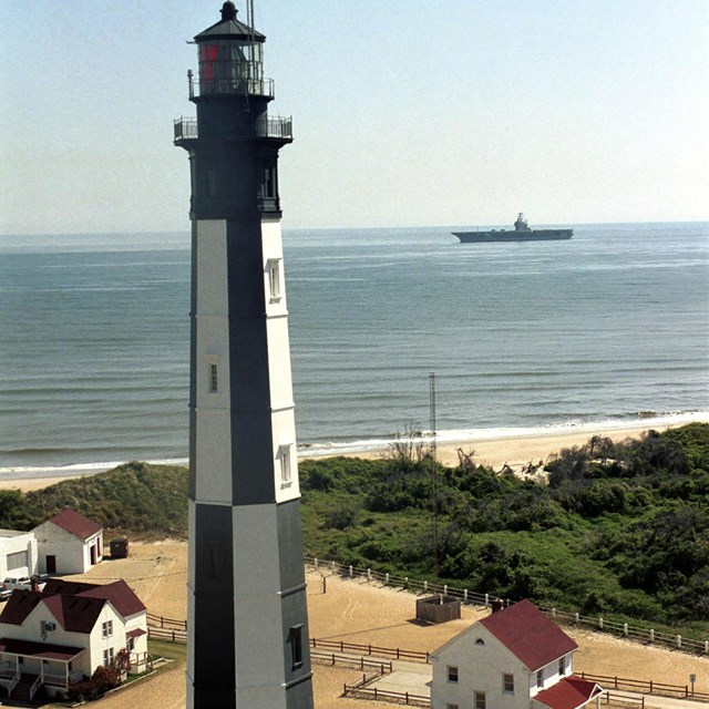 A yellow sandy beach overlooking the ocean. A black and white checked lighthouse dominates the shore