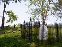A gated iron fence encloses a marble war memorial in the woods at the edge of a field.