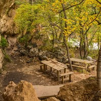 Stone tables and benches are located near a cliff with rock overhangs and alcoves.