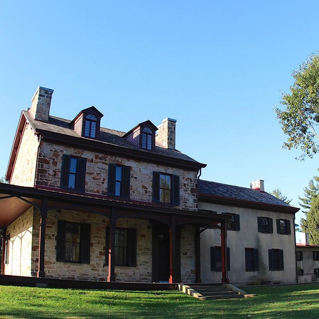 A large white stone house with wooden dormers. A grassy green area surounds it. A blue sky above.