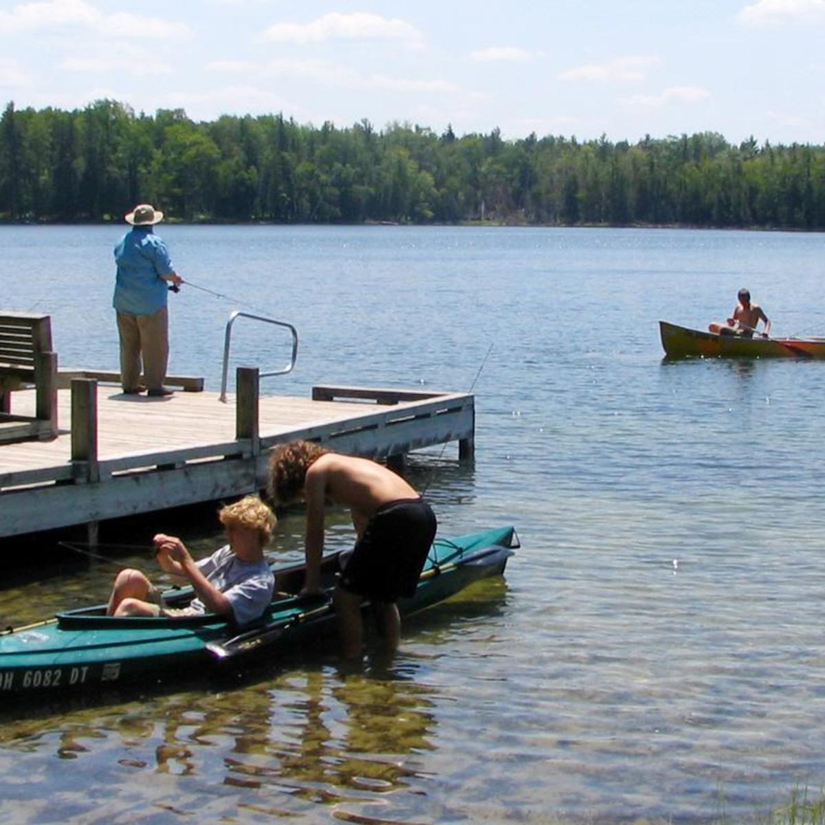 A woman fishes from the dock, canoeists paddle by, kayakers unload along the shore