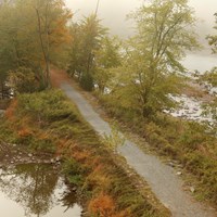Gravel path flanked by grass, trees. River on 1 side, remains of partially-filled canal on other.