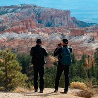 Two hikers pause along a path to view a red rock landscape of badlands and spires at a cliff\'s edge