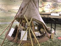 A museum exhibit of a Native American teepee, partial rolled up. Several small artifacts are shelter