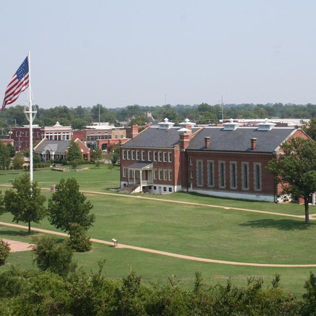 Aerial view of three-story red brick building on left next to tall flag pole and parade ground.