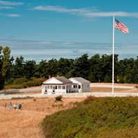 Historic white buildings in a grassy field. Hikers are going along various trails.