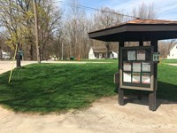 Information kiosk stands to the right of a gray trail that crosses a road to a small white house.