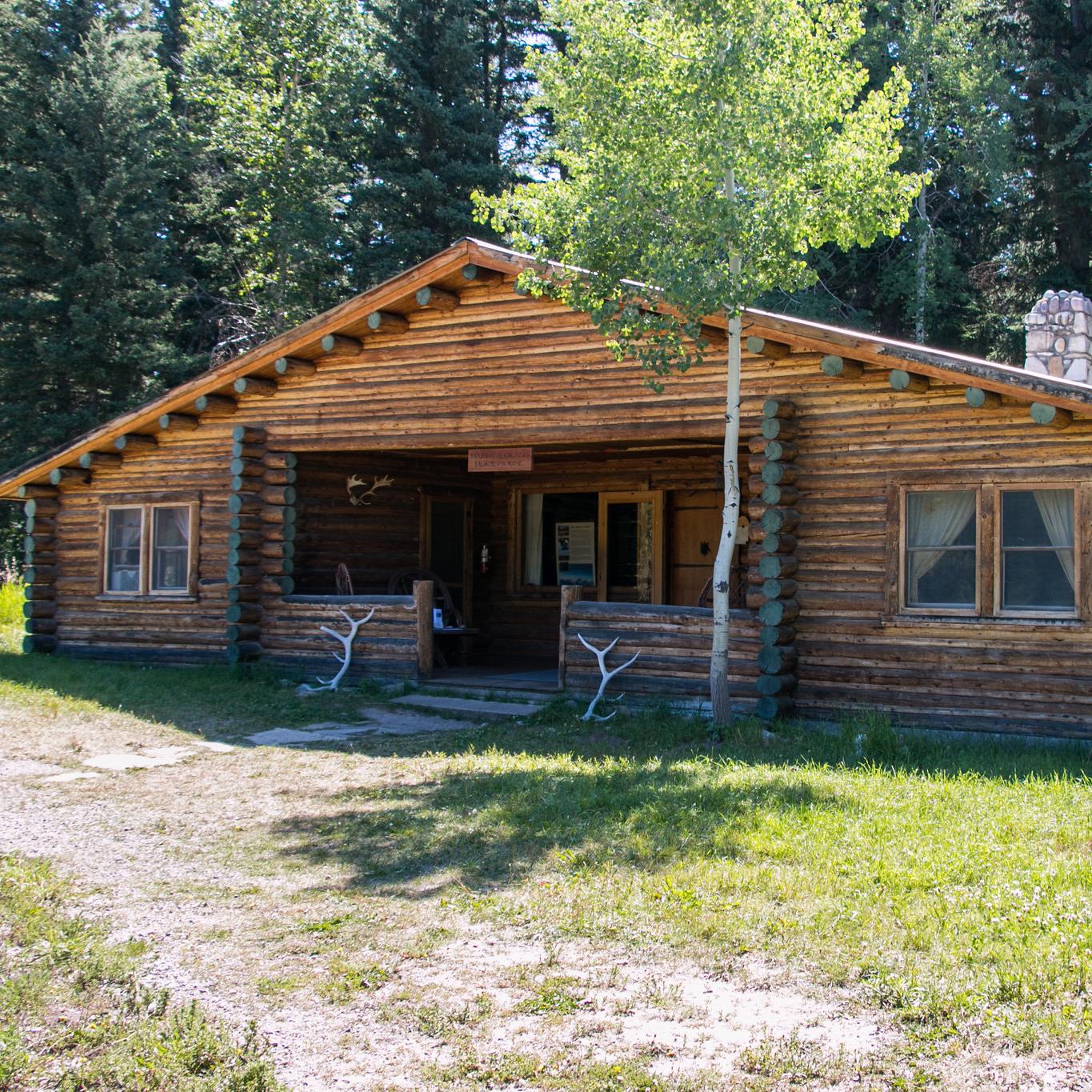 A brown, log cabin building with a river stone chimney and elk antlers resting on the front porch.