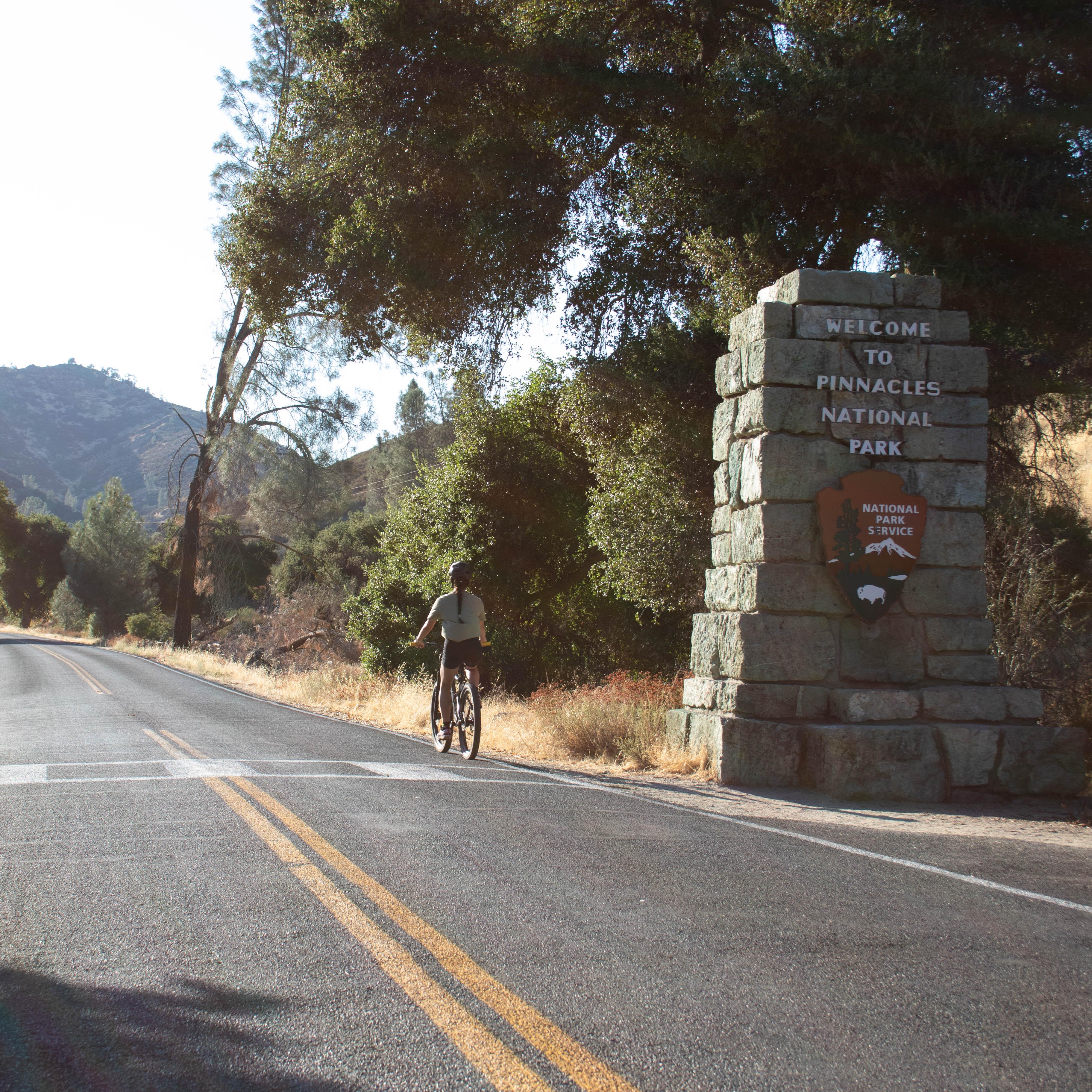 Visitor seen along a paved roadway riding a bicycle past a park service sign made into a rock pillar