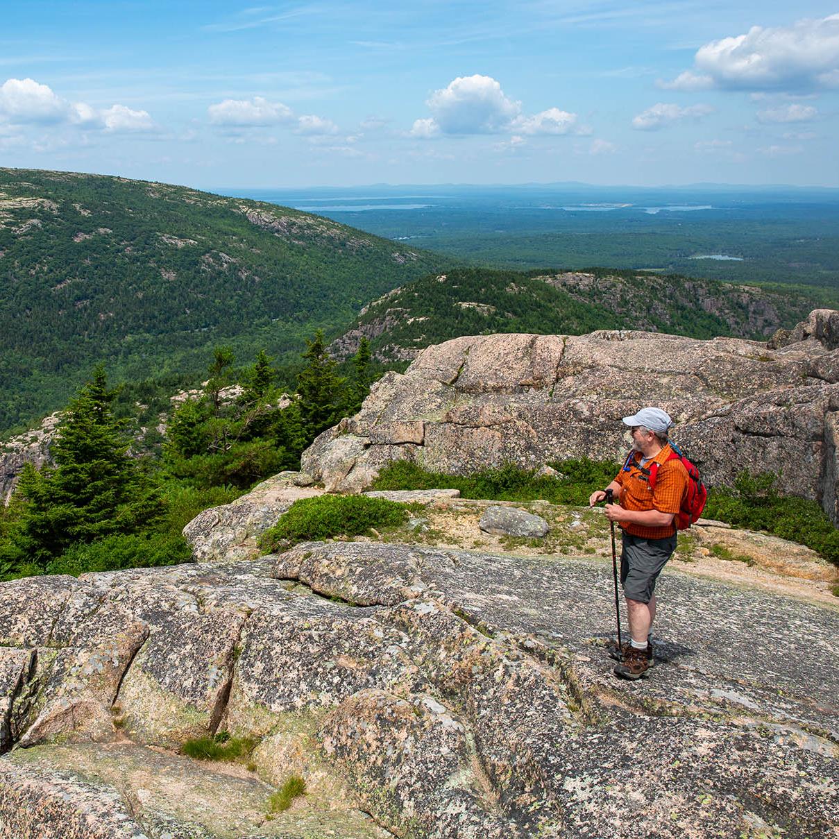 A hiker stands on an exposed granite peak, looking out at mountains rising across the valley.  