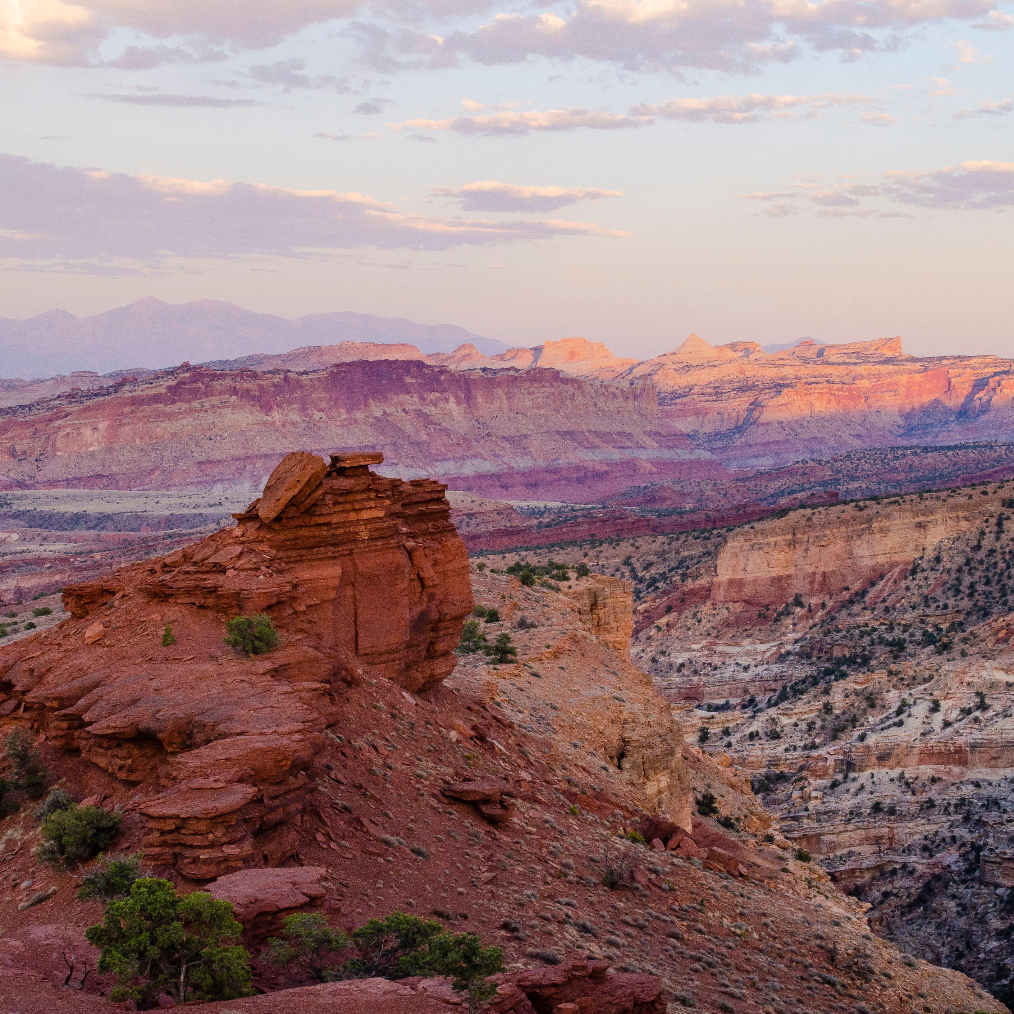 Colorful red rocks, striped cliffs, mountains in the distance, and a pale blue moving to purple sky.