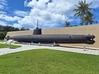 A large black submarine with a periscope resting on a concrete plinth in front of a concrete wall.