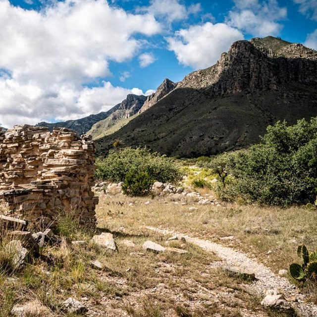 A ruin of an old stone wall, next to a trail that leads into distant mountains.