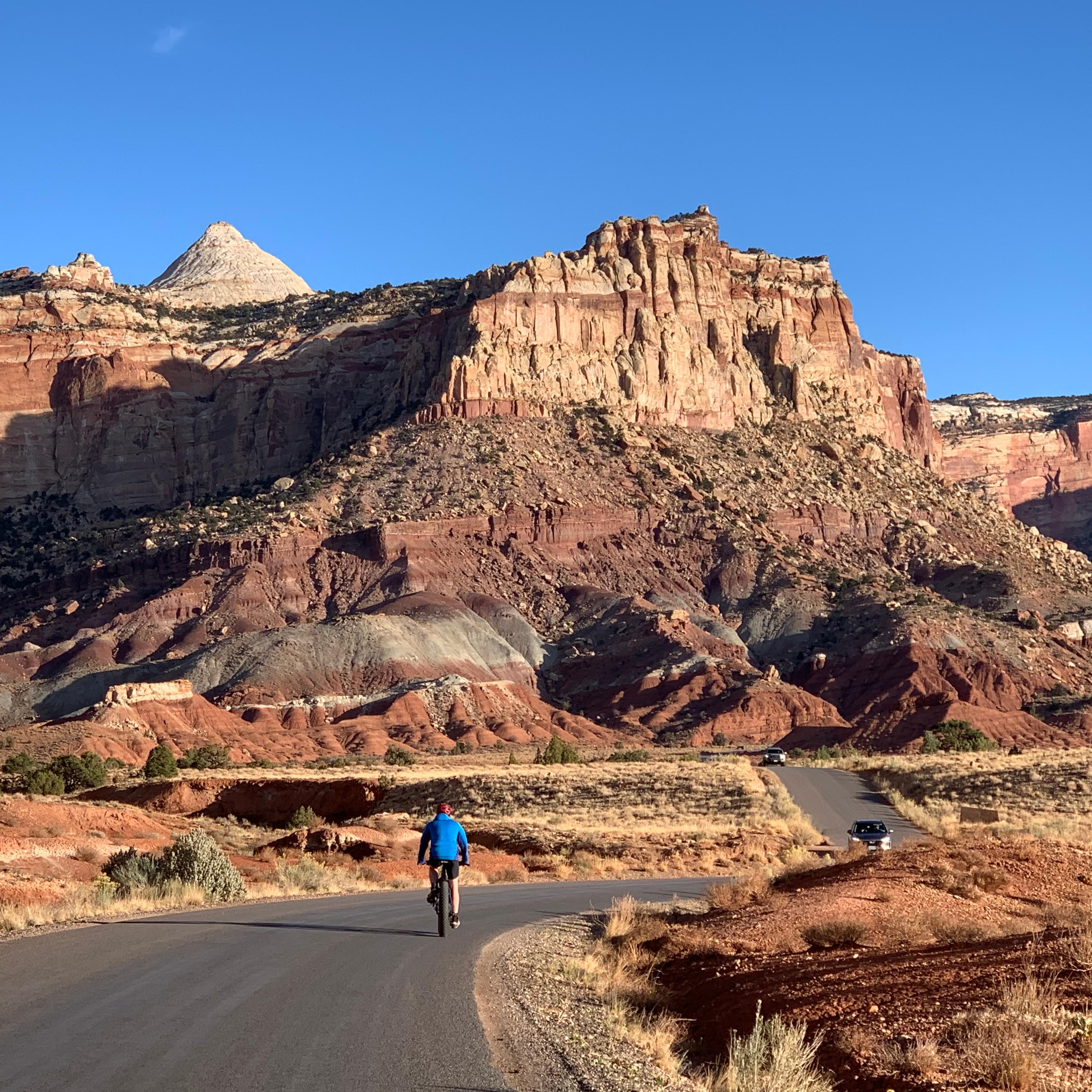 Person on bicycle on paved road with cars approaching and colorful cliffs above, and blue sky.