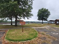 A brown sign in a grassy patch in a parking lot with distant trees in a park setting next to a river