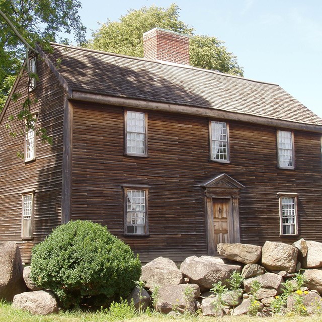 A large brown wooden house with five windows and one door on the front and a large chimney. 