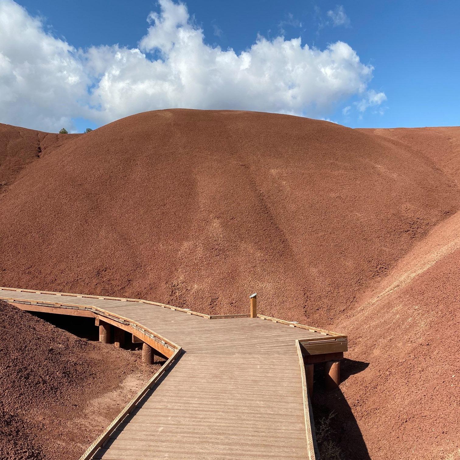 A wooden boardwalk goes between two red claystone hills. Cloudy blue skies are in the background.