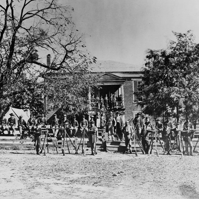 Black and white image of Union soldiers sitting on a split rail fence in front of a brick building. 