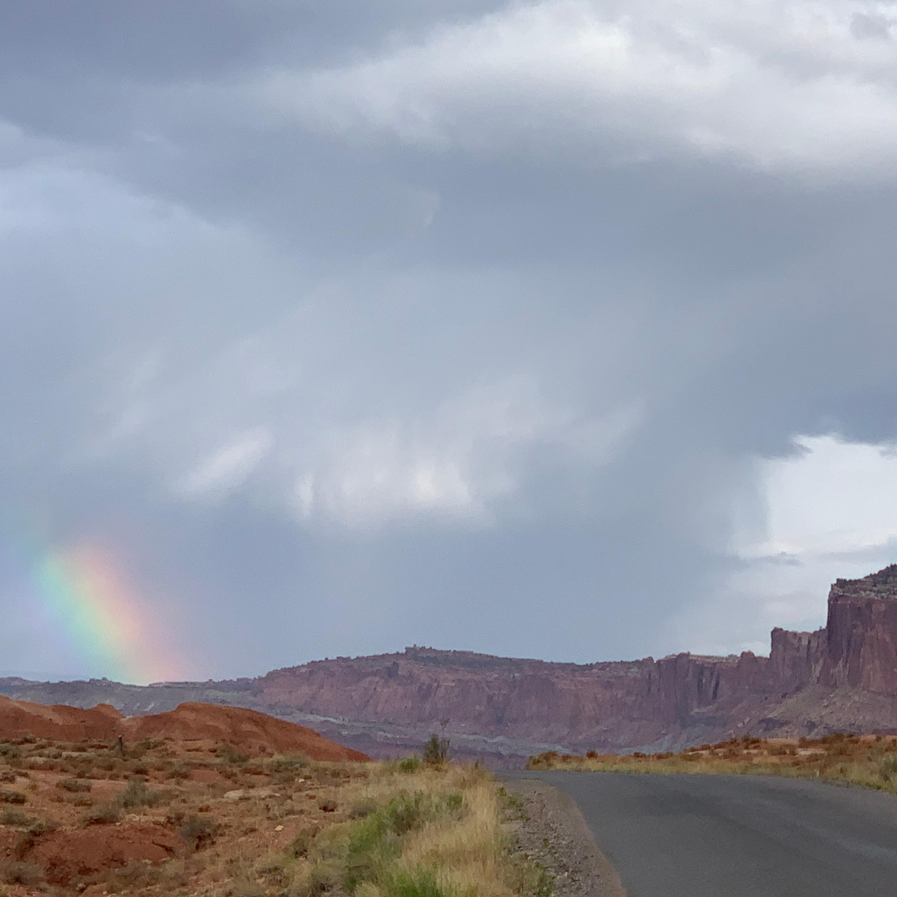 Stormy skies with a small spot of a rainbow, red cliffs, and a blacktop road. 