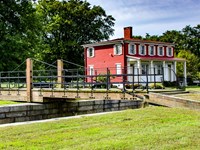 A red brick two-story building with a small wooden bridge over a stone canal, surrounded by greenery