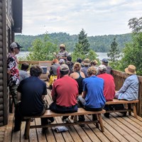 A park ranger gives a program to a group of people on a porch with Washington Harbor behind.