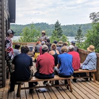A park ranger gives a program to a group of people on a porch with Washington Harbor behind. 