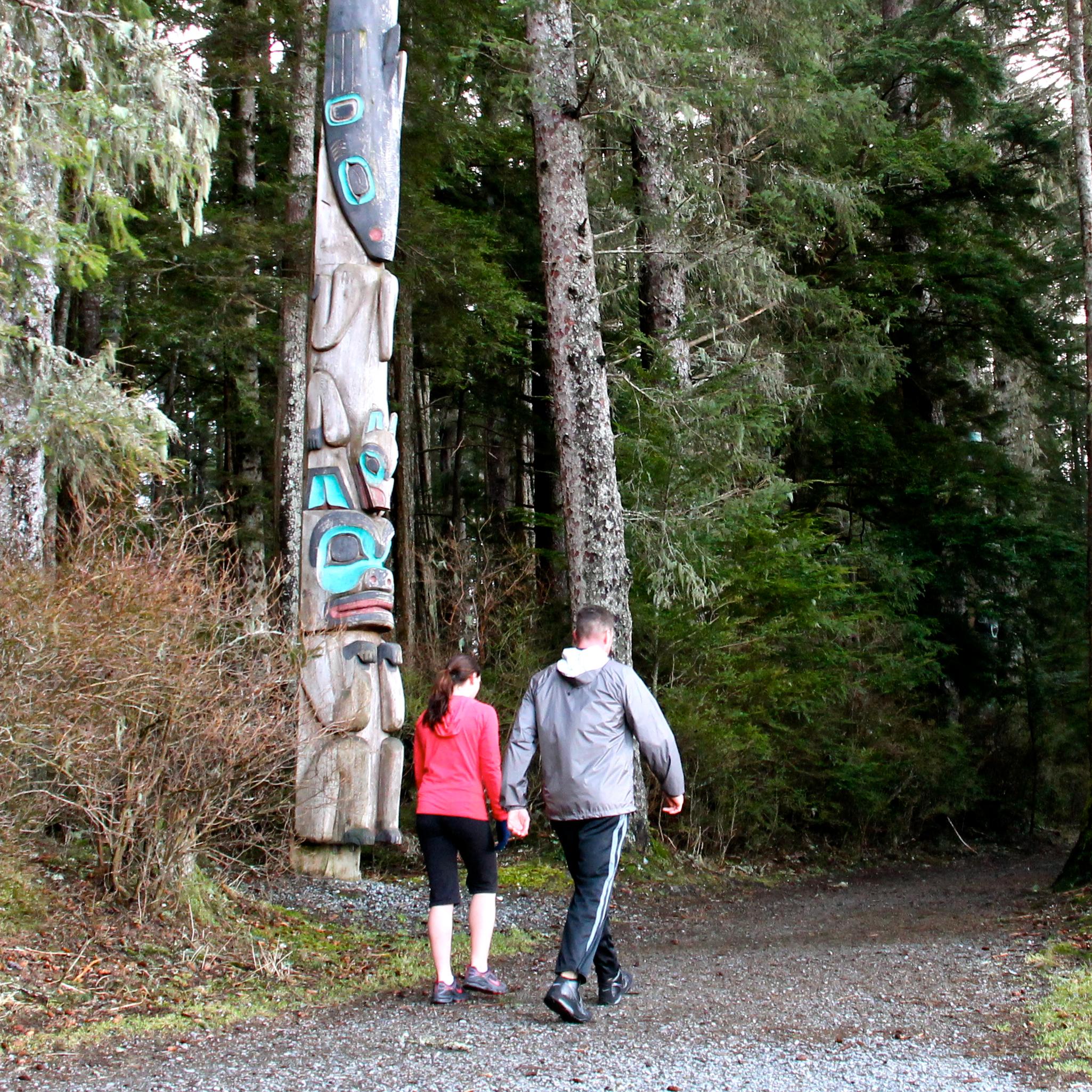 A man and woman walk by trees and a totem pole on a gravel path