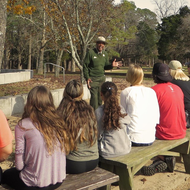 A row of people on a bench. You see their backs. They are sitting watching a park ranger speak. 
