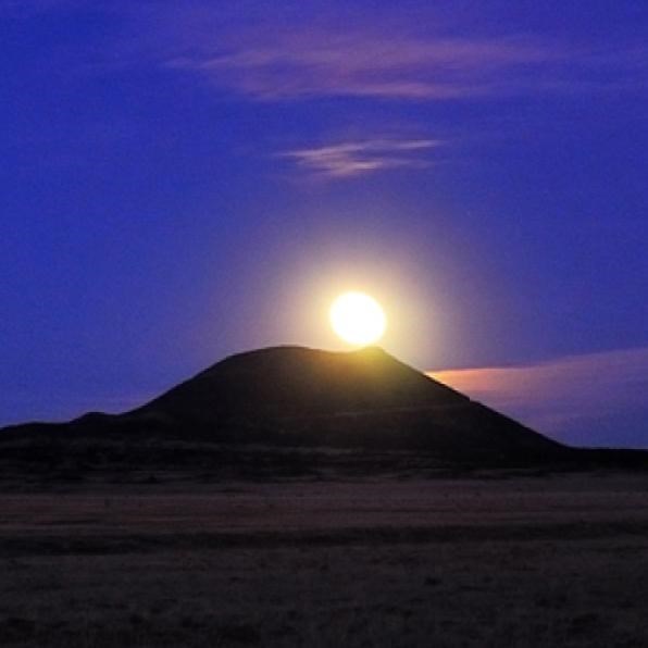 A bright moon rises over a cinder cone mountain at night. 