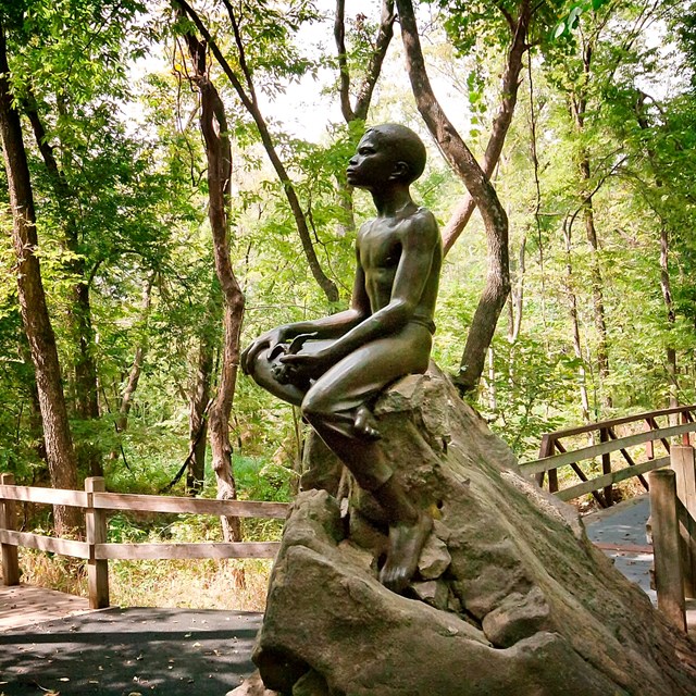 A sculpture of a young boy (George), sitting on a rock with a plant in is left hand. 