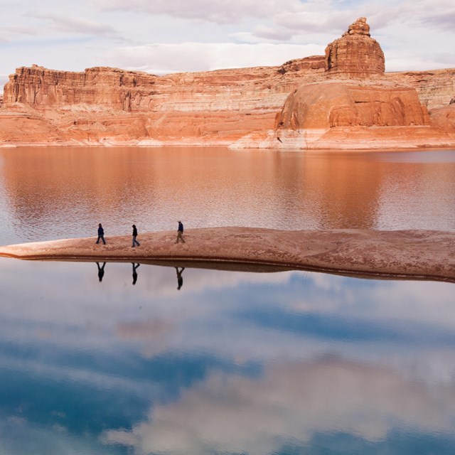 Three figures walk a thin rock outcropping surrounded by Lake Powell.