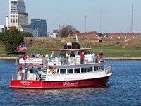 A red and white sightseeing boat with passengers near Fort McHenry and a city skyline in the backgro