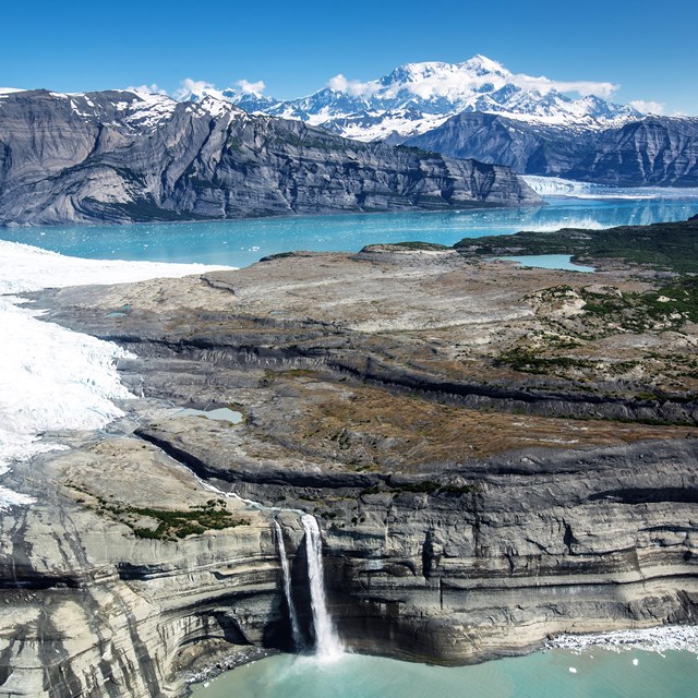 coastal bay with waterfall and glacier and large snow-covered mountains in the background