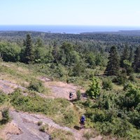 Two people with backpacks walk down a trail on a ridge surrounded by forest.