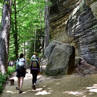 Two people walk side by side next to colorful rock cliffs; each carries a baby on their back.