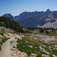 A trail meanders through a rocky, alpine meadow with jagged peaks in the background.