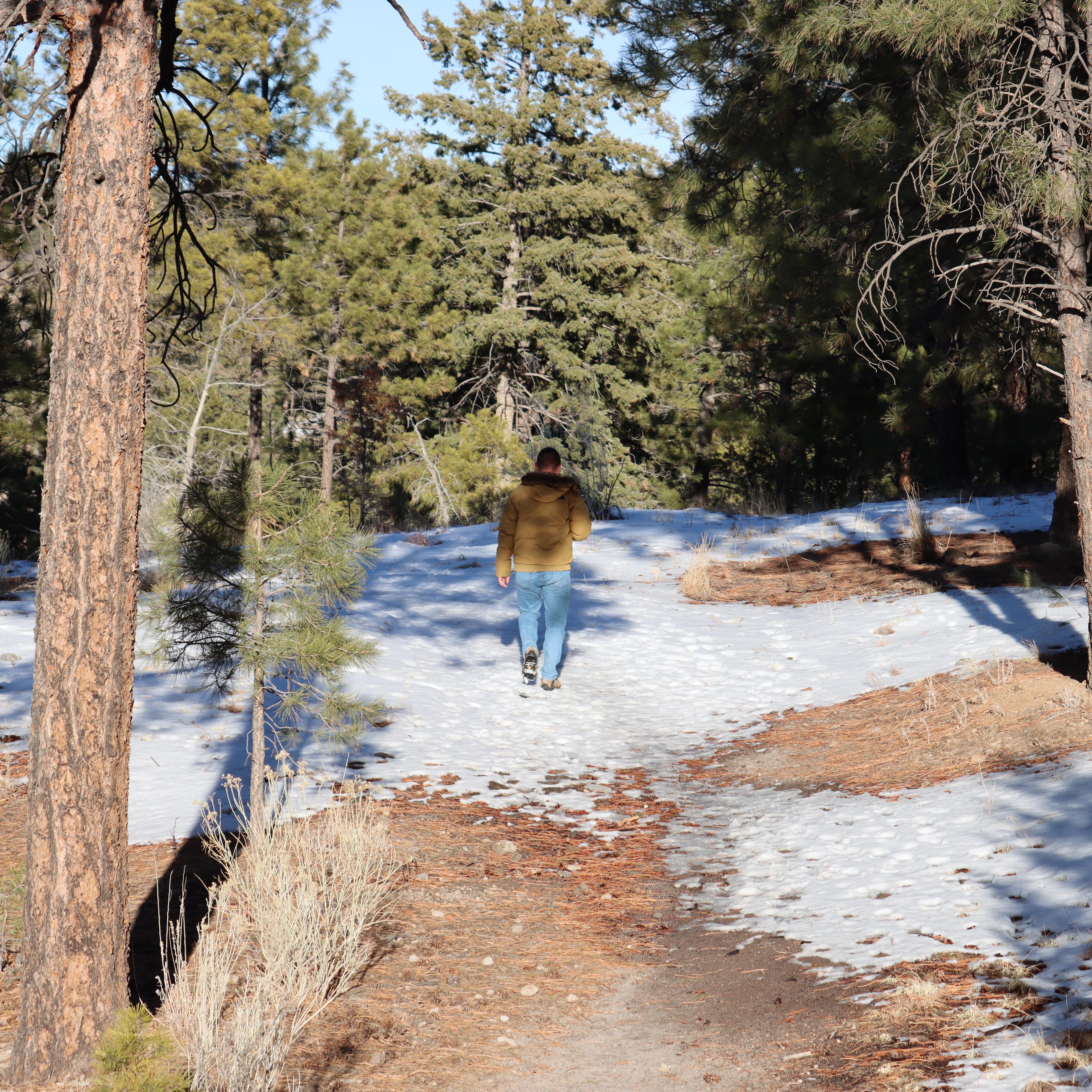 A person in a tan jacket and jeans walks on a snowy trail surrounded by trees.