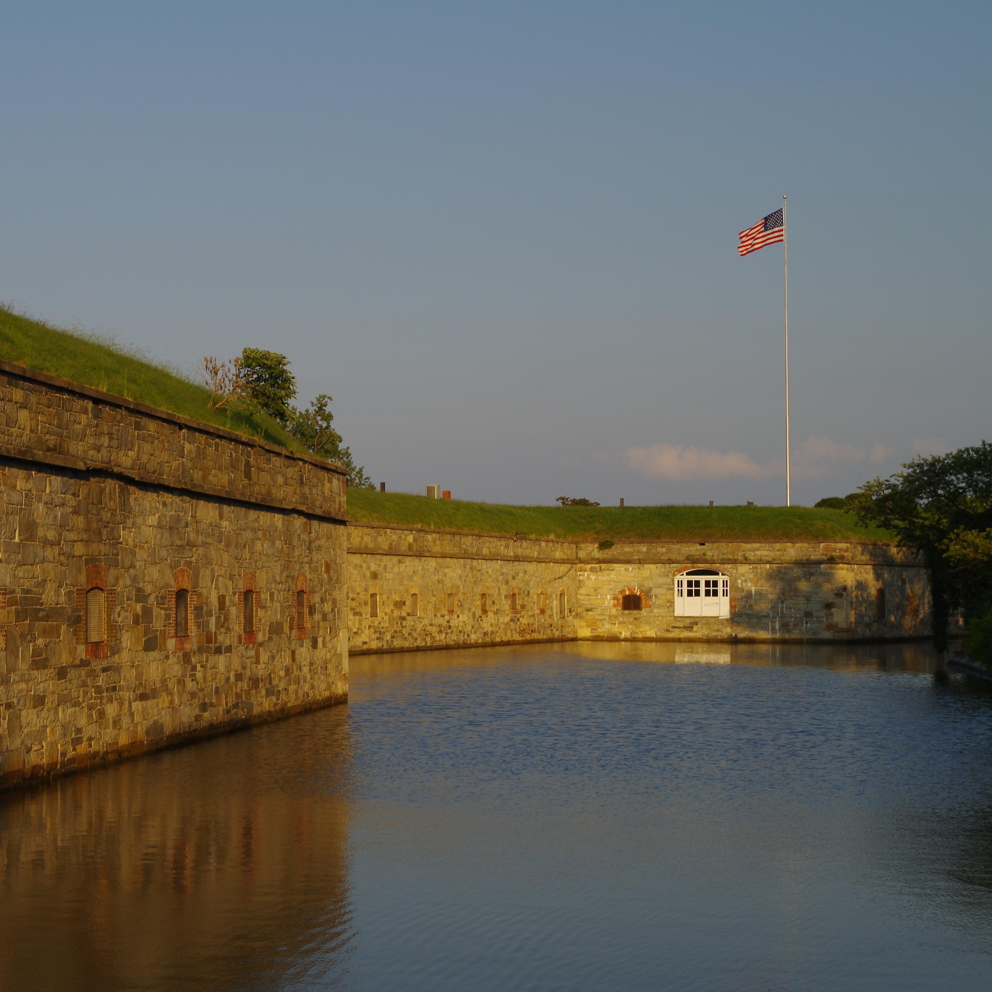 The setting sun lights a stone fort wall where a US flag flies on a high staff. 