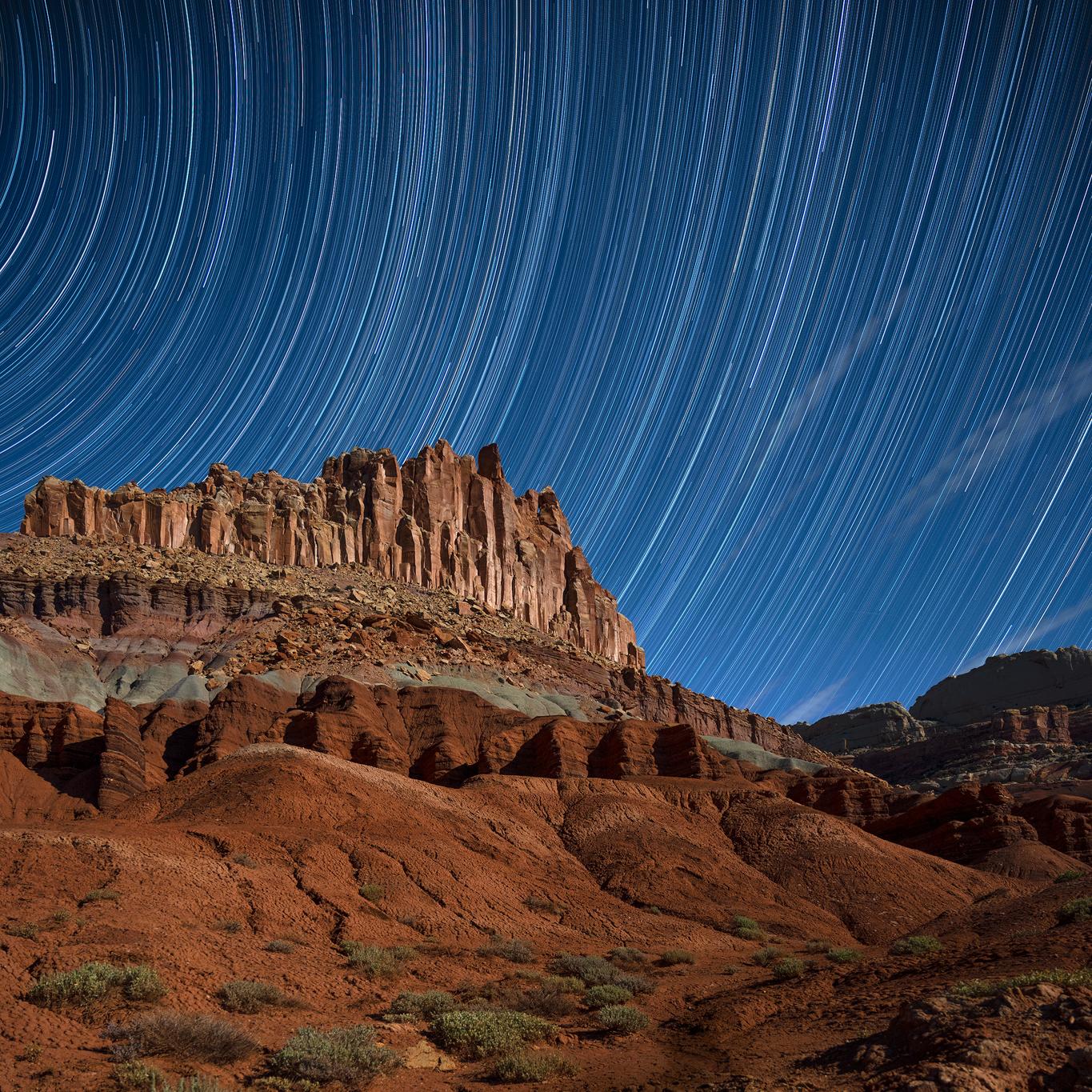 White lines representing circular star trails in dark blue sky, above colorful red cliffs. 