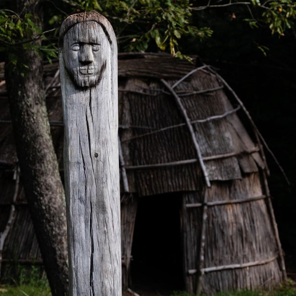 A Native American recreated town has a longhouse structure and sculpture with human face